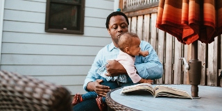 Man holds a baby at a picnic table in a backyard.
