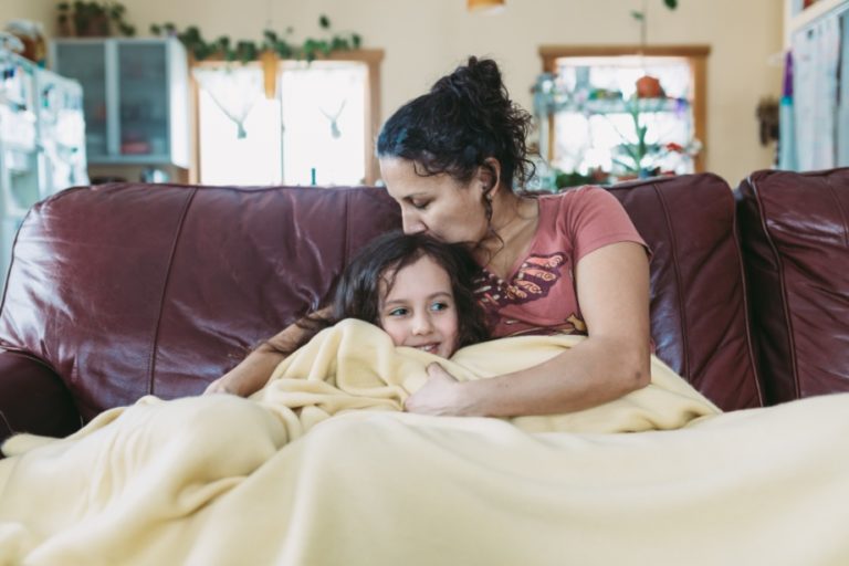 A woman snuggles with a little girl under a blanket on a sofa.